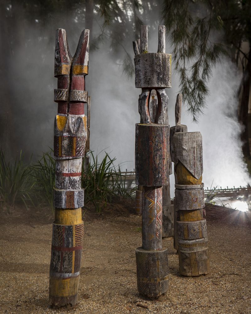 Three carved poles painted in red, yellow, and white ochre with mist behind