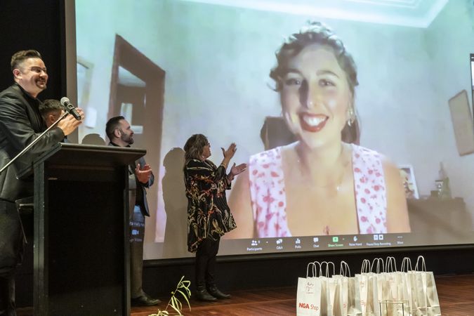 Three people are on stage clapping, while a large projection on the stage of a video call shows a person on screen smiling
