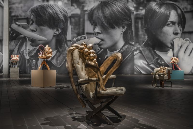 In a large gallery space sits a golden sculpture of a female form with many breasts sitting on a chair
