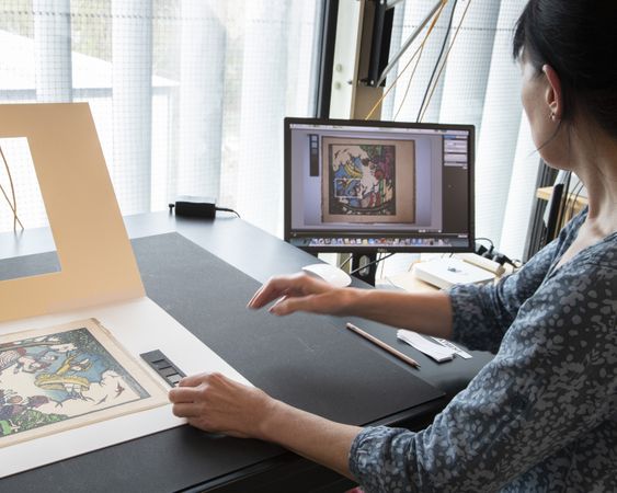 A woman sits at a desk cross-checking a work of art on paper with an image on a computer screen
