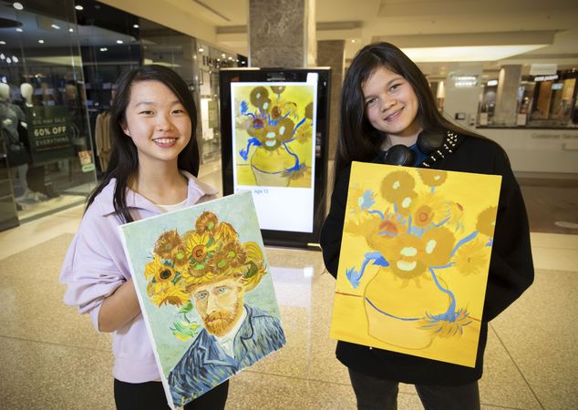 Two children stand in a shopping mall holding paintings they have done representing Van Gogh's Sunflowers