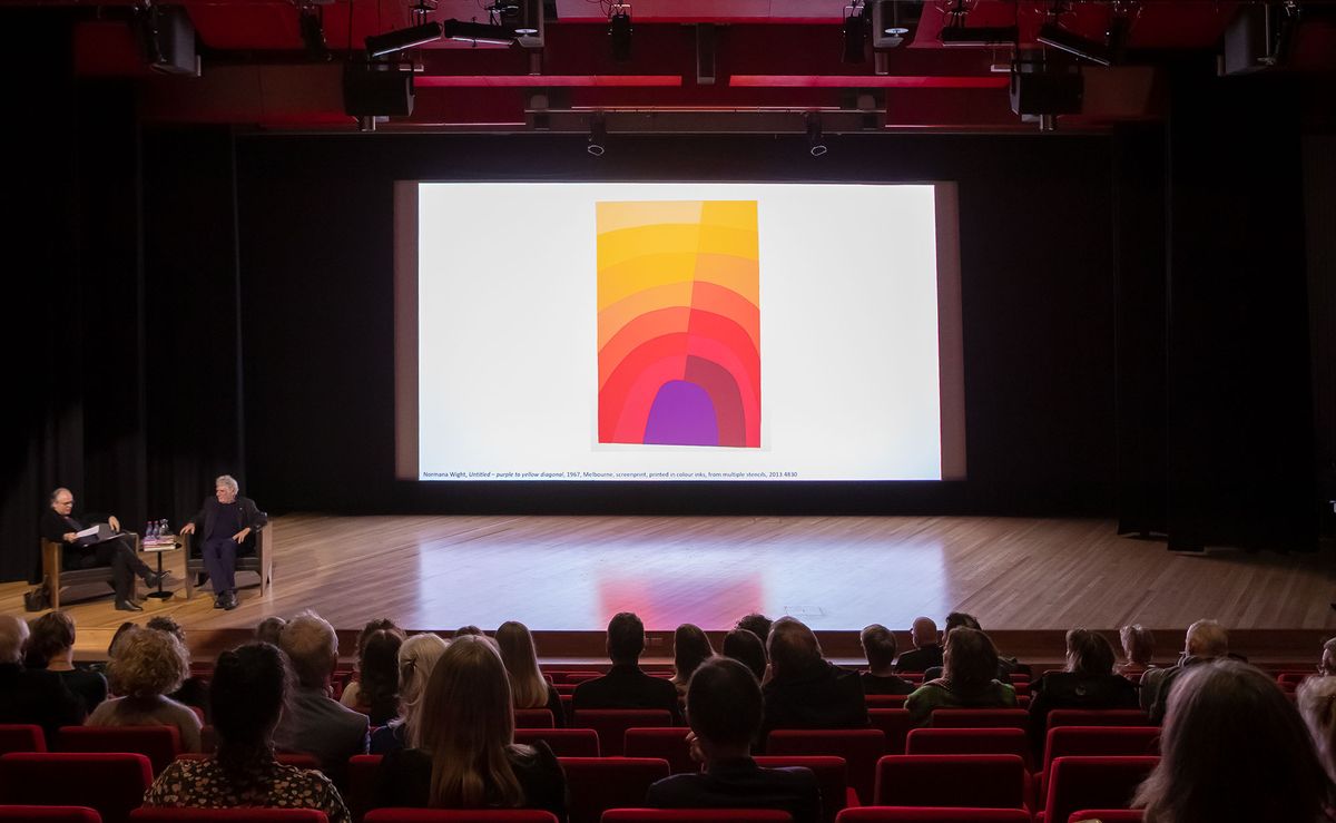 People sit in a darkened theatre watching two people in conversation on stage to the side of pictures on a large screen