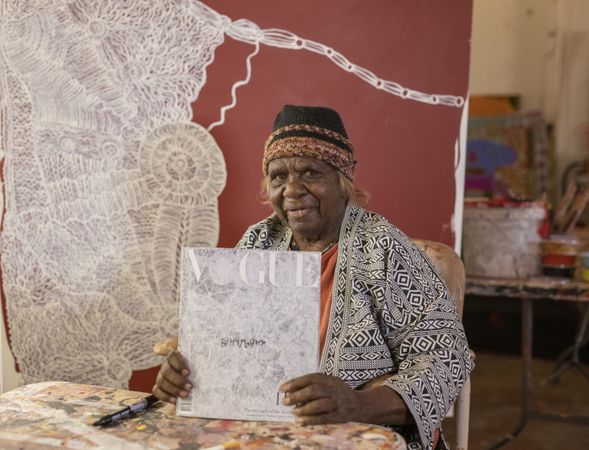 An older woman sits in front of her painting, holding a copy of Vogue magazine that has her artwork on the cover