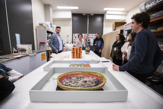 Indigenous art objects are sitting on a large table in a conservation lab. People are standing around the table, one woman is speaking to the group.