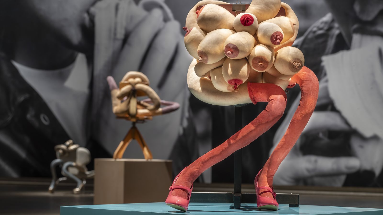 Installation view of a sculpture of a human-like form made up of a cluster of many breasts and two long legs with long pink socks, sitting on a chair