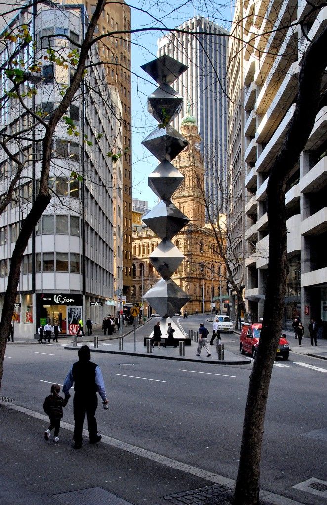 A large-scale sculptural work of seven steel trapezoidal prisms stacked vertically, and surrounded by buildings.