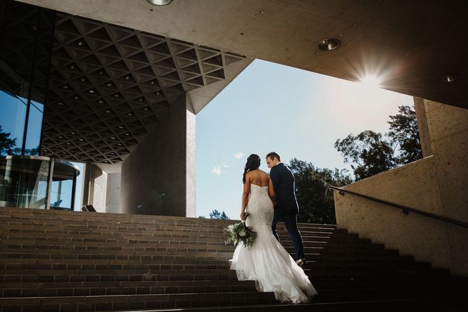 A couple in wedding attire facing away from the camera hols hands while walking up the stairs outside a large building
