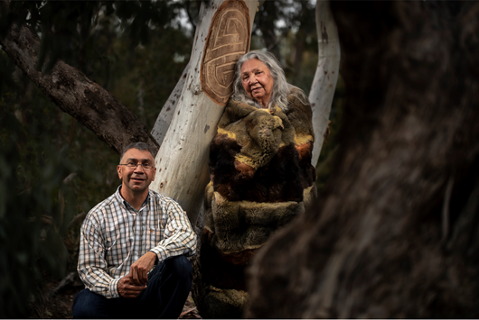Two First Nations artists standing next to a scarred tree work of art