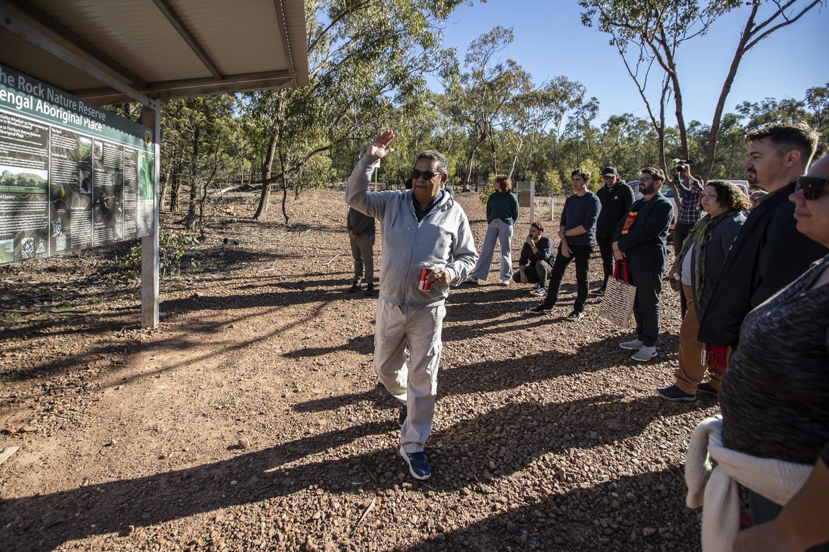 Outside a man in a grey tracksuit is talking to a group of people gathered around him