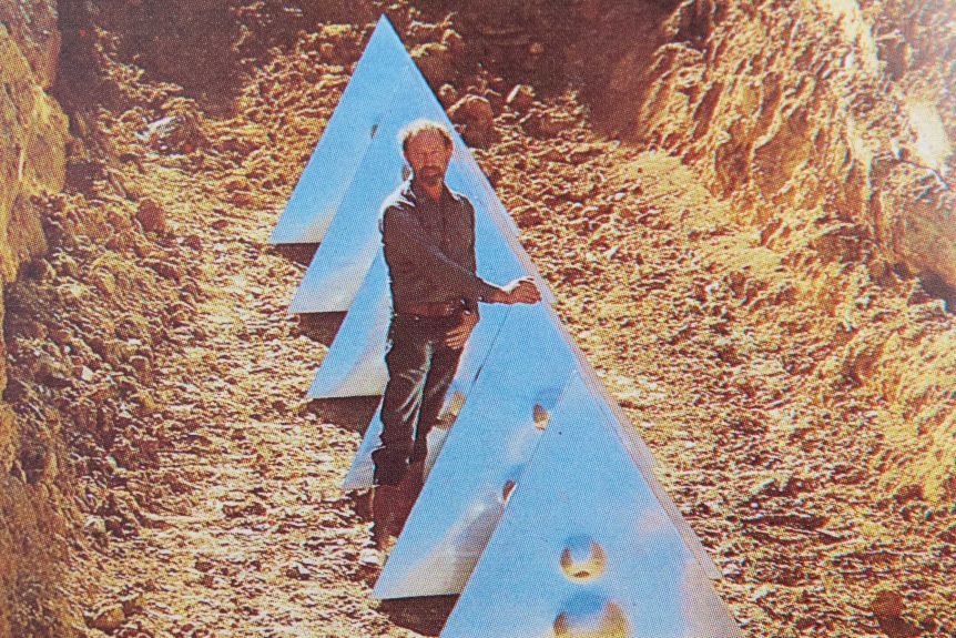 A young Bert Flugelman stands in a dug out trench alongside six large, silver triangles