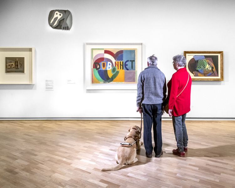 Photograph of two visitors facing artworks with companion dog by side