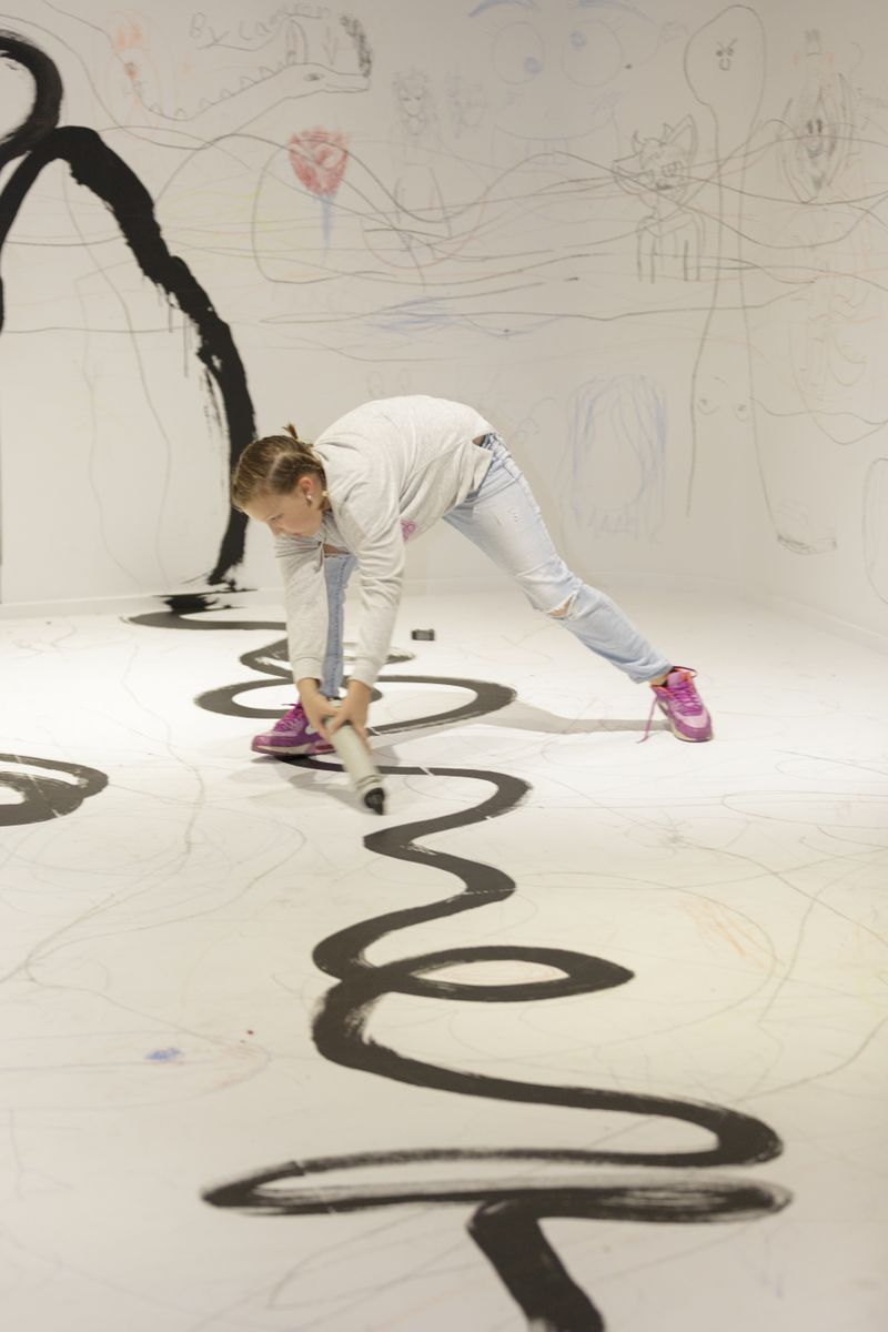In a room with drawing marks and scribbles all over the white floor and walls child is drawing on the floor using an oversize crayon