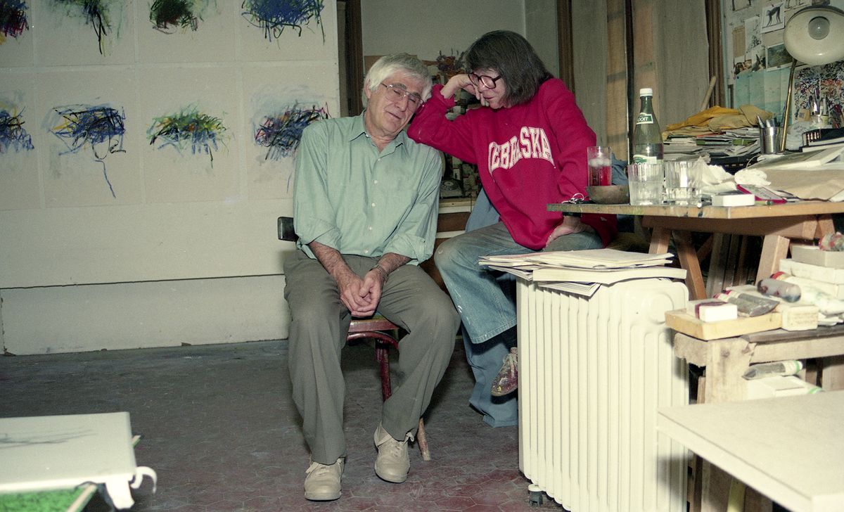 A photograph of Joan Mitchell and Kenneth Tyler sitting on chairs in an artist studio.