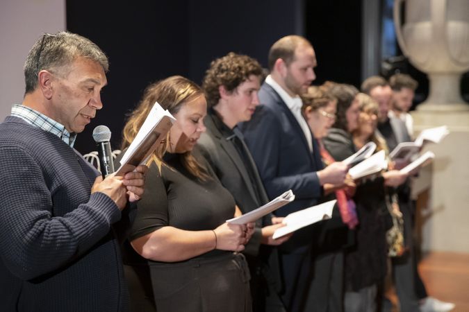 People are standing in a line holding microphones and reading/singing together from a booklet