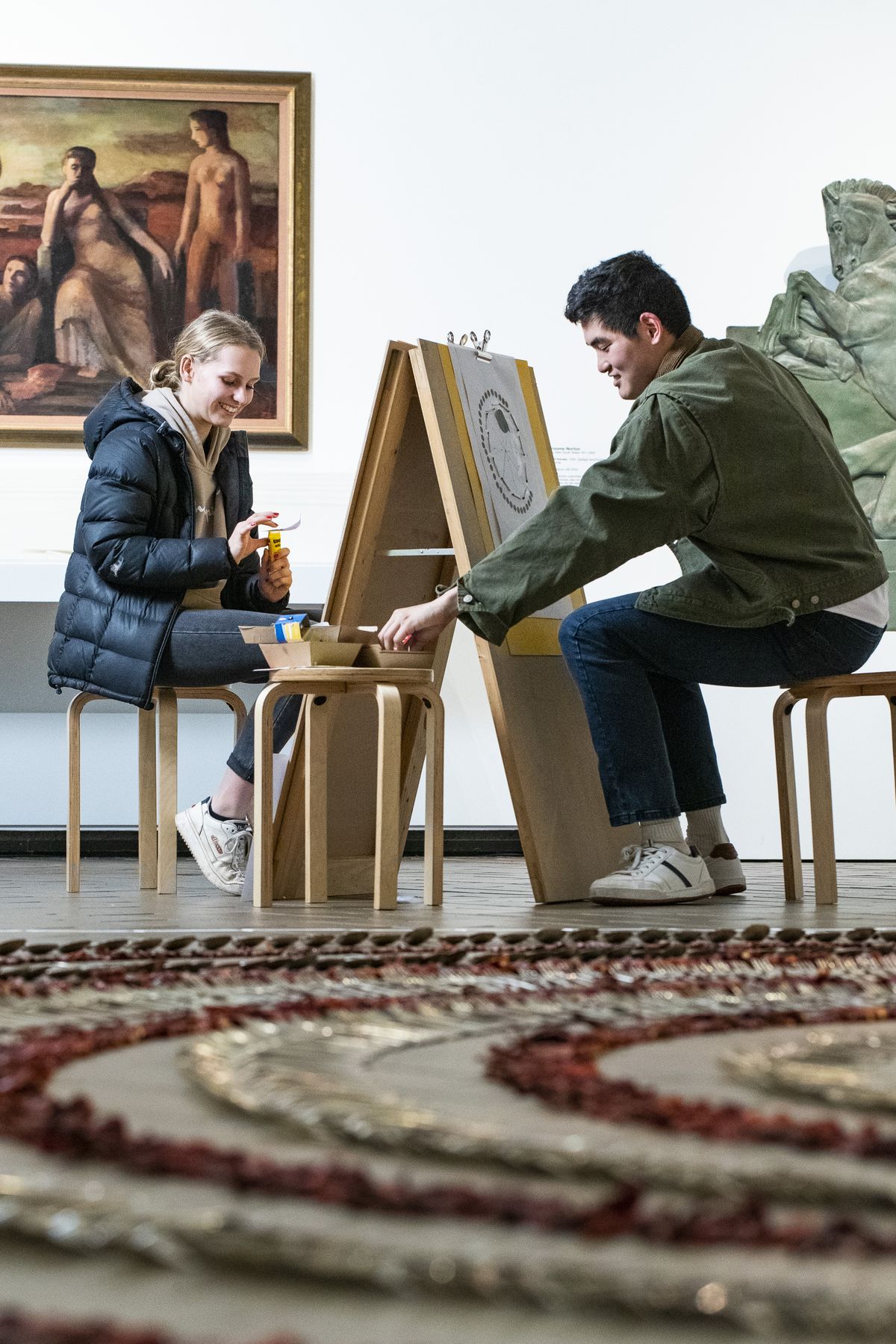 Two young people sit either side of an easel in a gallery space