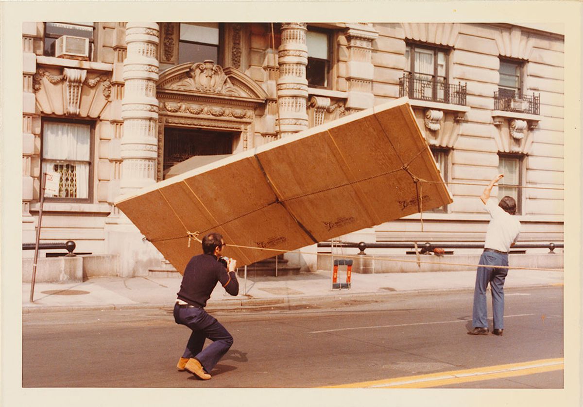 Two men using a pulley system to lower a boxed painting from a tall apartment block