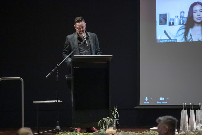 A man is standing behind a lectern on a stage, smiling as he give a speech. There is a projection of people joining via video call displayed on the stage beside him.