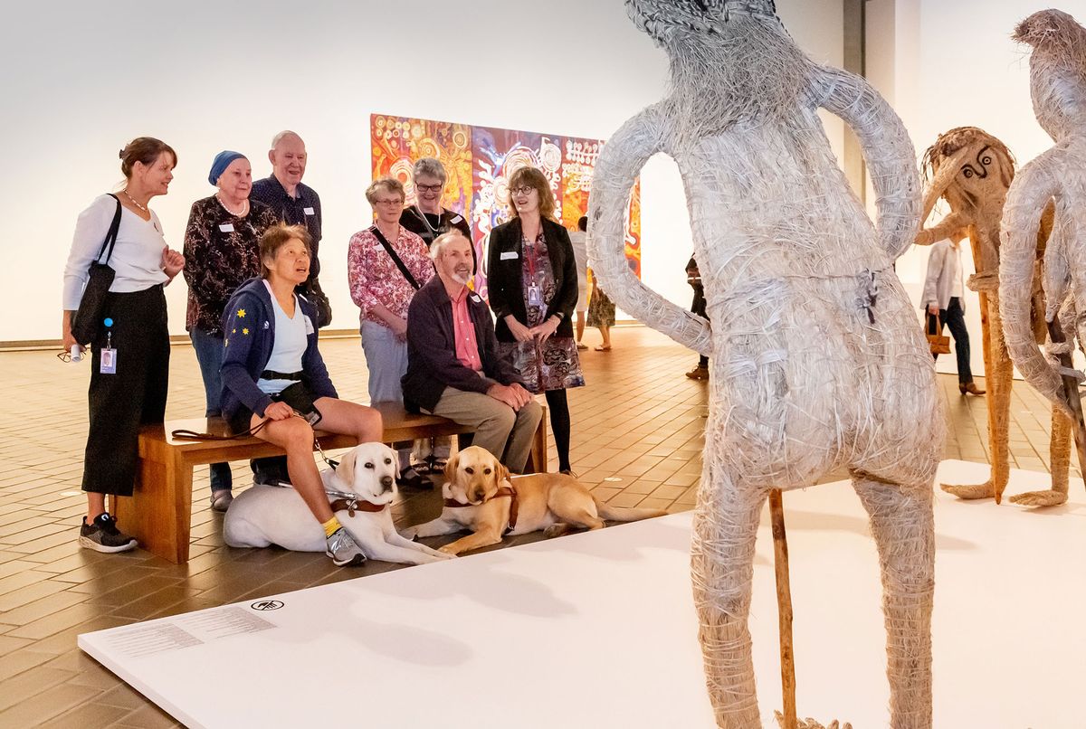 A group of people and two guide dogs sit looking at sculptures of humans woven with native grasses