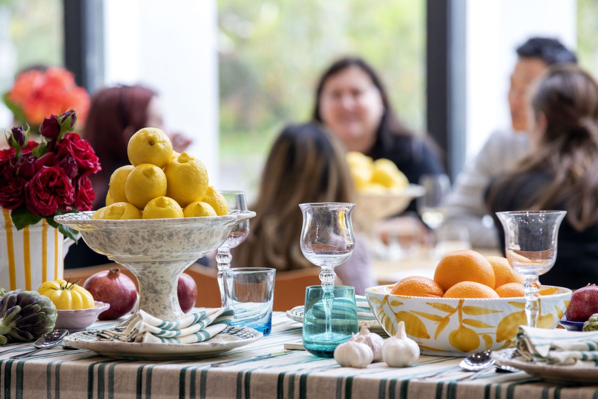 A table with a bowl with oranges and a bowl with lemons, garlic, onions and other vegetables and a vase of Roses and wine and drinking glasses and cutlery, with a table of people in the background