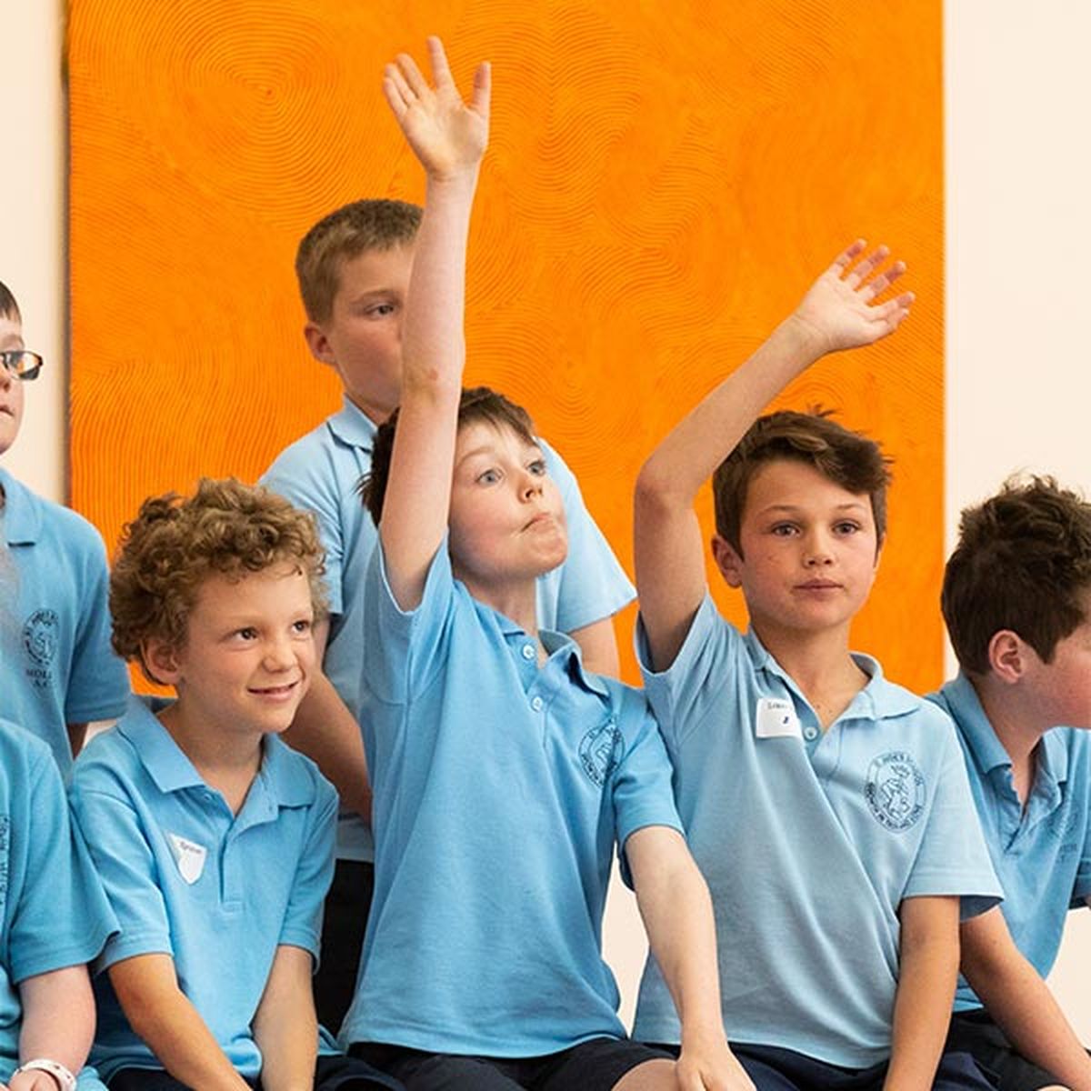 A seated group a primary aged school boys in school uniforms and two boys raising their hands up in the air