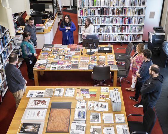 Birds-eye view of two large tables in a library, surrounded by book shelves. Each table has archival material laid out on it, and people are standing around the tables