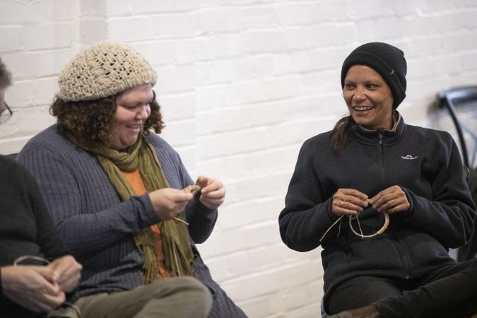Two women are sitting next to each other talking and laughing as they weave with natural grasses