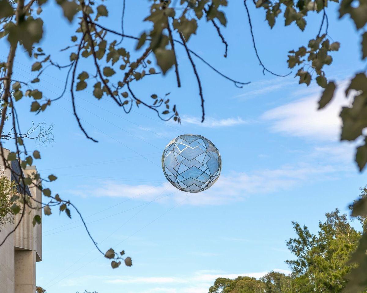 A large spherical sculpture hanging from large wires in the sky is visible through a gap in tree branches