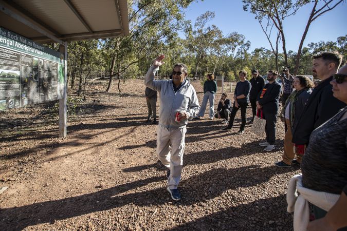 Outside a man in a grey tracksuit is talking to a group of people gathered around him