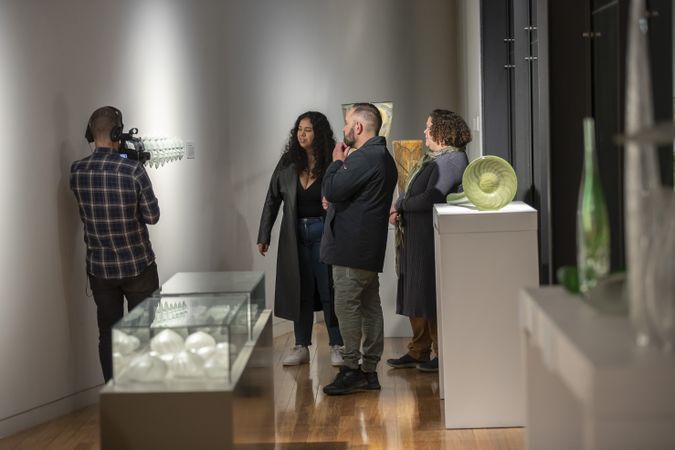 Three people are standing in a gallery looking at works of art while one person with a video camera films the art